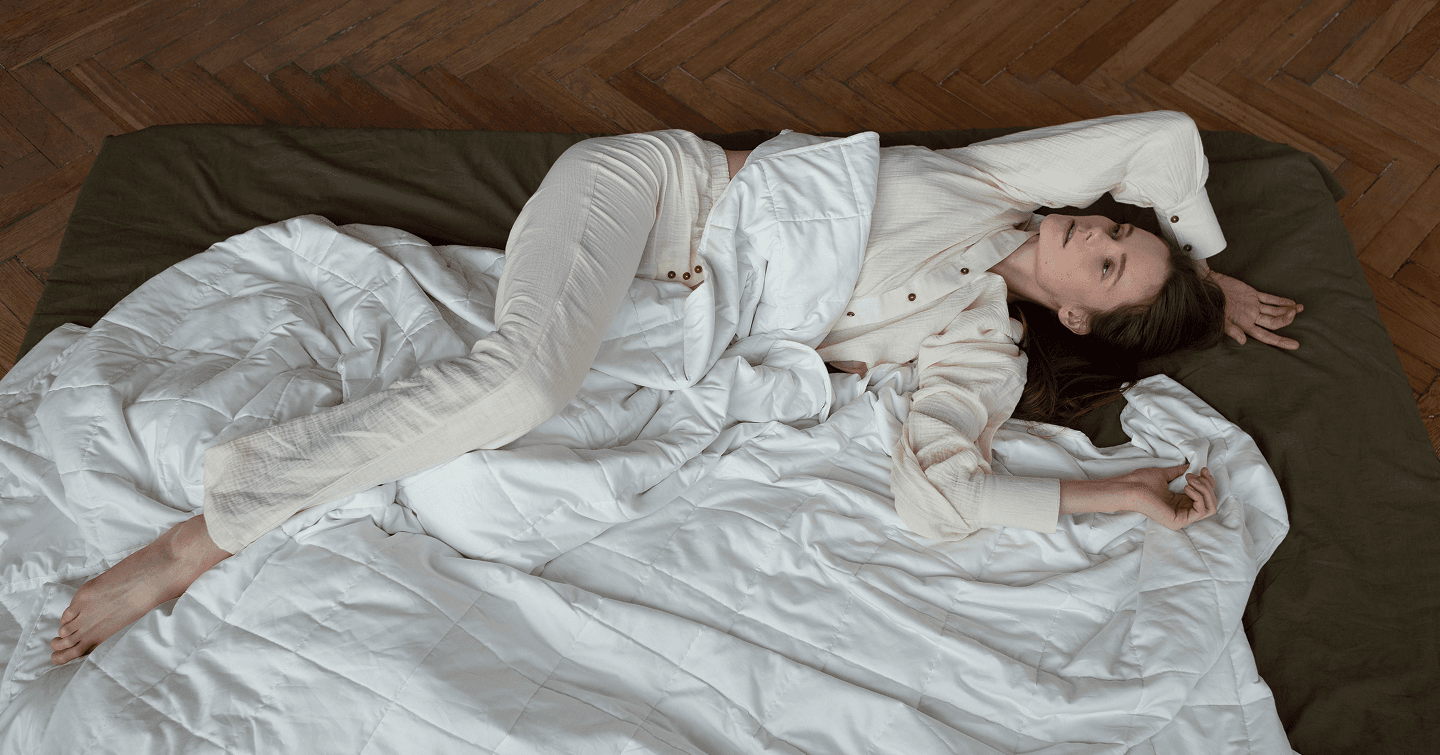 A woman resting on a bed layered with a white quilted weighted blanket over an olive green sheet, set on a herringbone wood floor.