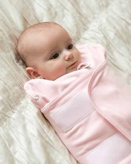 Baby lying on a bed swaddled in a pink Lullaboo sleeping bag with hook-and-loop closures, looking toward the camera