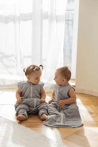 Two babies sitting on the floor in grey knit sleeping bags near a sunlit window