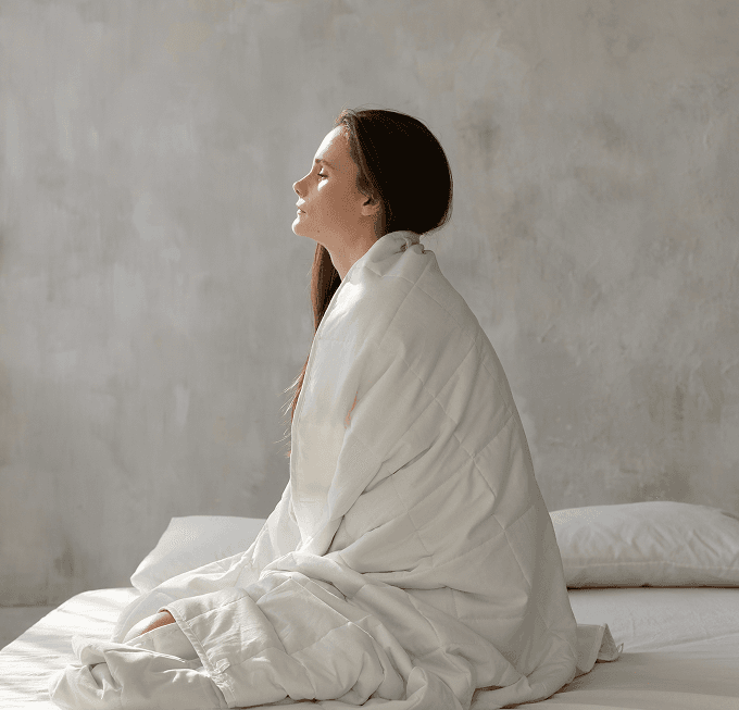 Woman sitting on a bed with her eyes closed, wrapped in a white Lullaboo weighted blanket