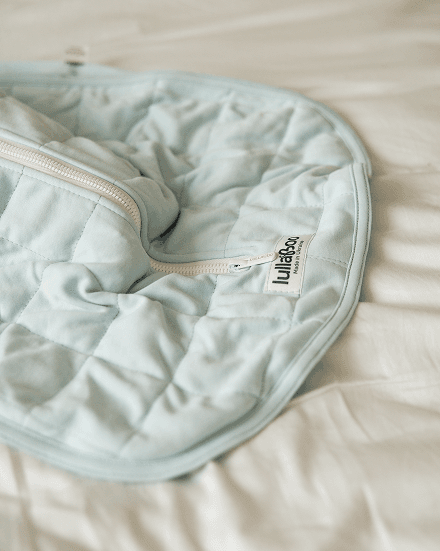 Close-up of an unzipped light blue quilted Lullaboo sleeping bag with its woven label, laid open on a bed