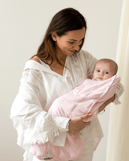 Mother in a white blouse smiling down at her baby, who is swaddled in a pink Lullaboo sleeping bag