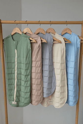 Five Lullaboo quilted sleeping bags in different colors hanging on a wooden clothing rack