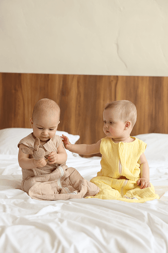 Two babies sitting together on a bed wearing tan and yellow sleeping bags